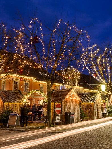 A festively decorated street with glowing trees and small wooden huts. The evening sky is dark blue, which highlights the lights even more. | © TV Thermen- & Vulkanland