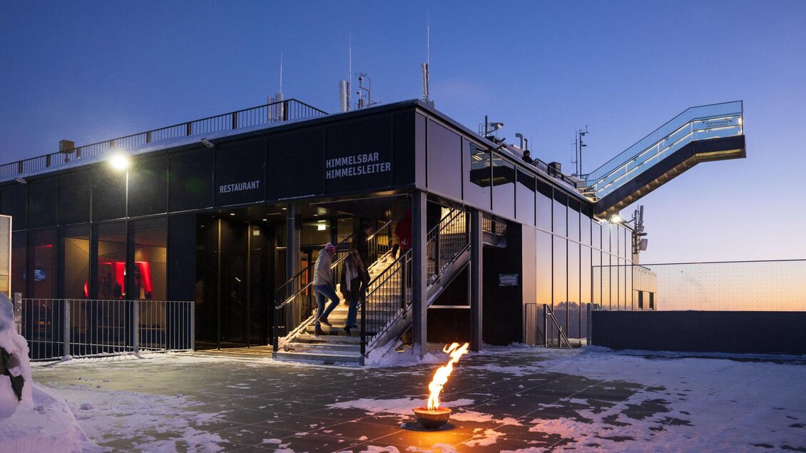 Ein modernes Gebäude mit Treppen und großen Fenstern, beleuchtet vor einem Sonnenuntergang. Im Vordergrund brennt eine kleine Flamme auf dem schneebedeckten Boden. | © Martin Huber