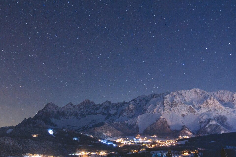 Ein klarer Nachthimmel mit vielen Sternen über schneebedeckten Bergen. Im Tal leuchten die Lichter eines kleinen Dorfes. | © Michael Simonlehner