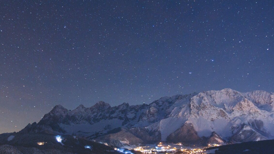 Ein klarer Nachthimmel mit vielen Sternen über schneebedeckten Bergen. Im Tal leuchten die Lichter eines kleinen Dorfes. | © Michael Simonlehner