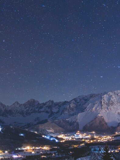A clear night sky with many stars above snow-covered mountains. In the valley, the lights of a small village shine. | © Michael Simonlehner