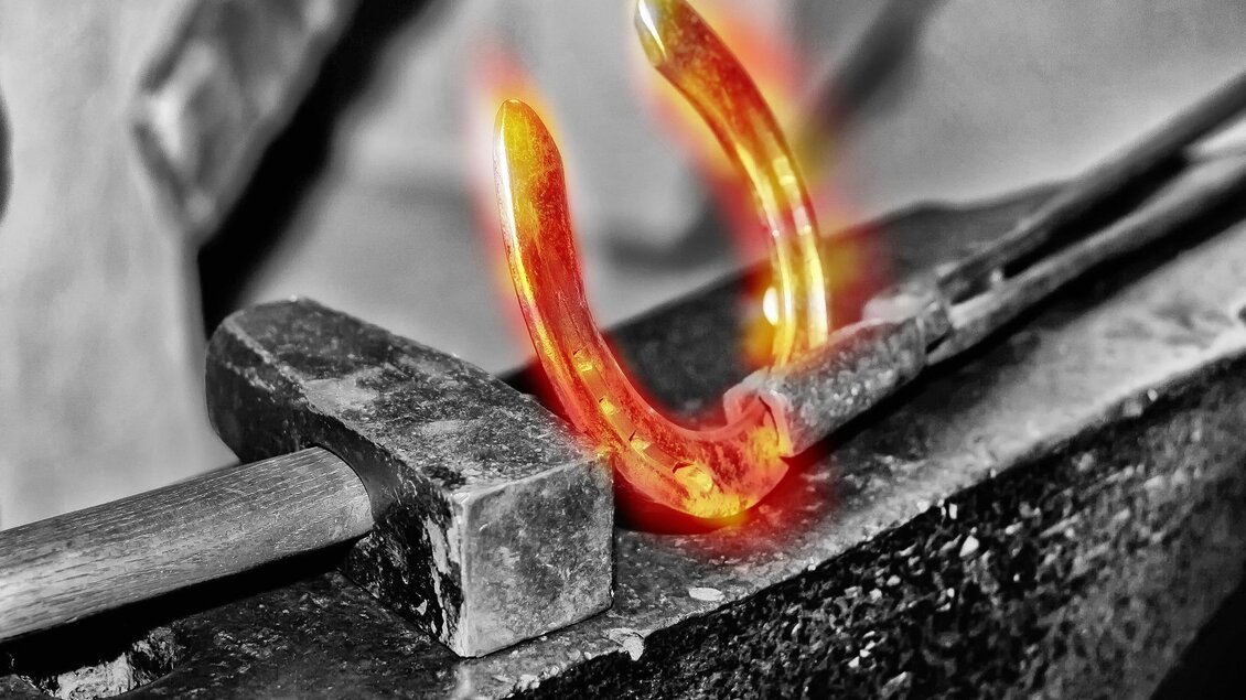 Ein glühendes Hufeisen liegt auf einem Amboss. Der Hintergrund ist schwarz-weiß, während das Hufeisen leuchtet. | © Pixabay Symbolfoto