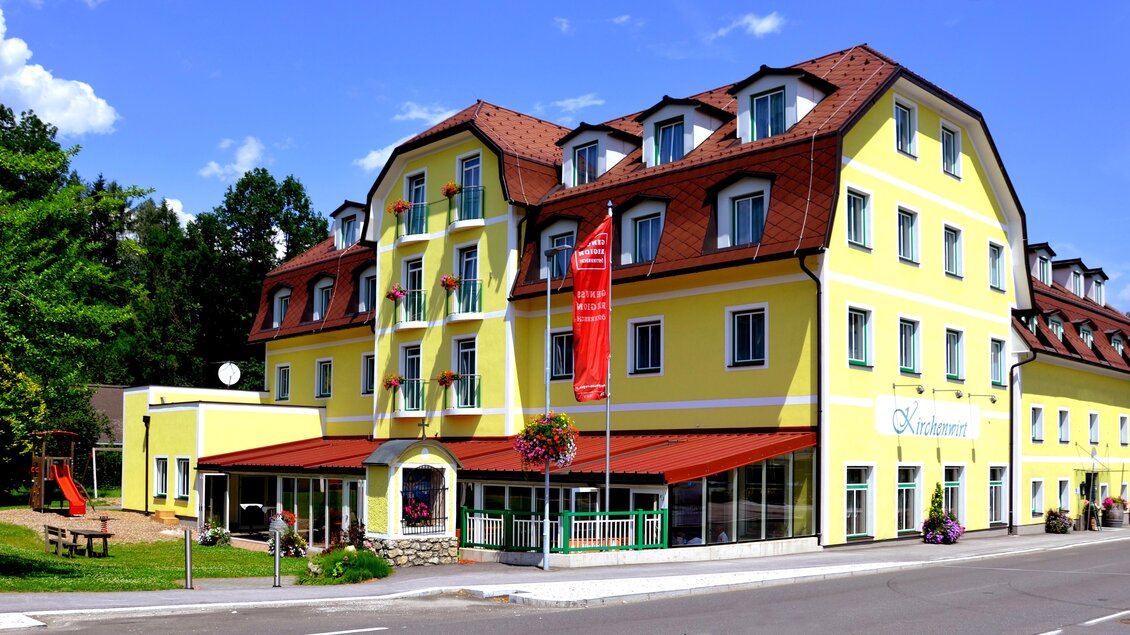 Ein großes gelbes Hotel mit mehreren Fenstern und einem roten Dach. Vor dem Gebäude gibt es eine kleine Grünfläche und Parkmöglichkeiten. | © Kirchenwirt Aigen