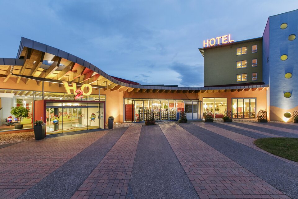 Ein modernes Hotelgebäude mit einer einladenden Fassade und hellen Lichtern. Der Eingangsbereich ist großzügig und freundlich gestaltet.