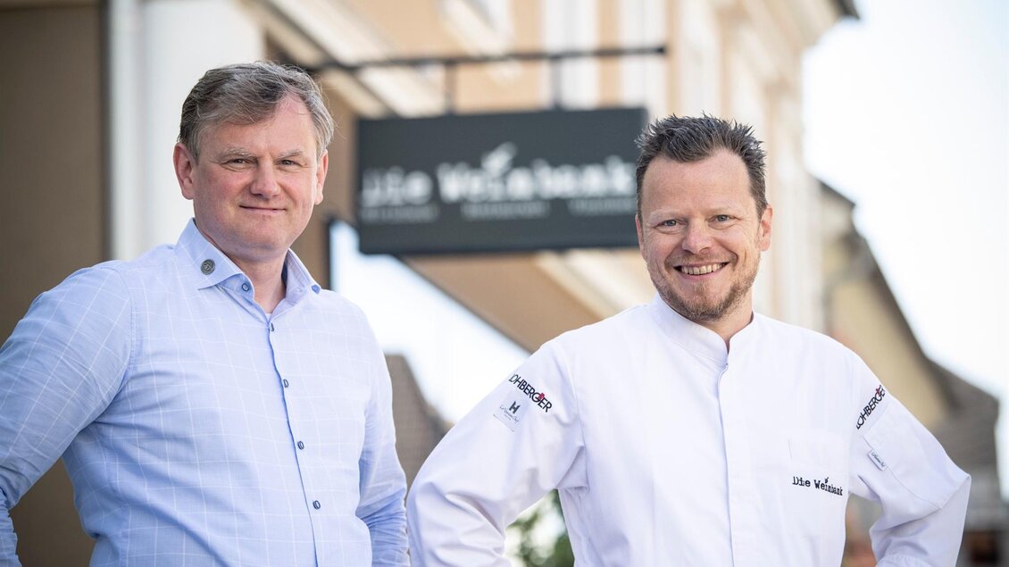 Zwei Männer stehen vor einem Restaurant mit dem Namen „Die Wildküche“. Der Mann links trägt ein blaues Hemd, der Mann rechts ist im Kochanzug gekleidet. | © Christian Jungwirth