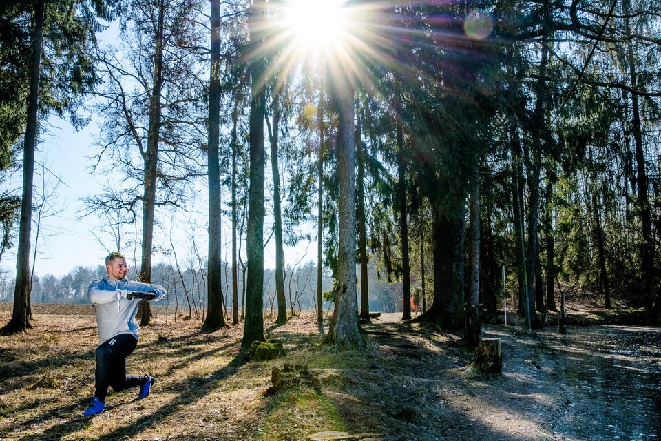 Ein Mann dehnt sich im Wald unter strahlendem Sonnenlicht. Die Umgebung ist grün und ruhig, ideal für sportliche Aktivitäten. | © TV Region Graz-Mias Photoart