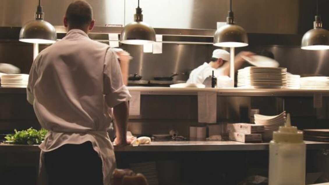 Ein Koch steht in einer belebten Küche und bereitet Gerichte vor. Im Hintergrund sind weitere Köche bei der Arbeit zu sehen. | © Bergmann