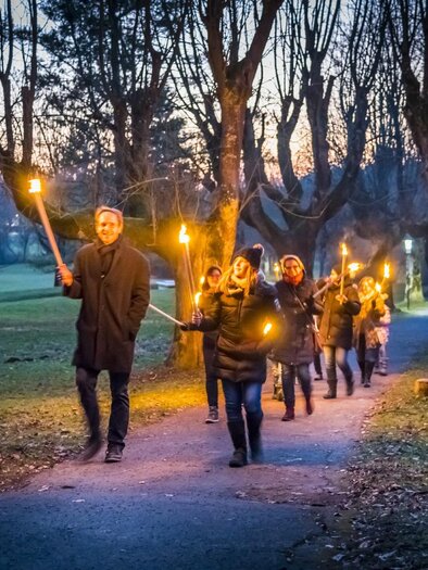 Eine Gruppe von Menschen geht nachts mit Fackeln durch einen Park. Die Bäume sind im Hintergrund sichtbar und es herrscht eine mystische Atmosphäre. | © Thermen- & Vulkanland, Werner Krug