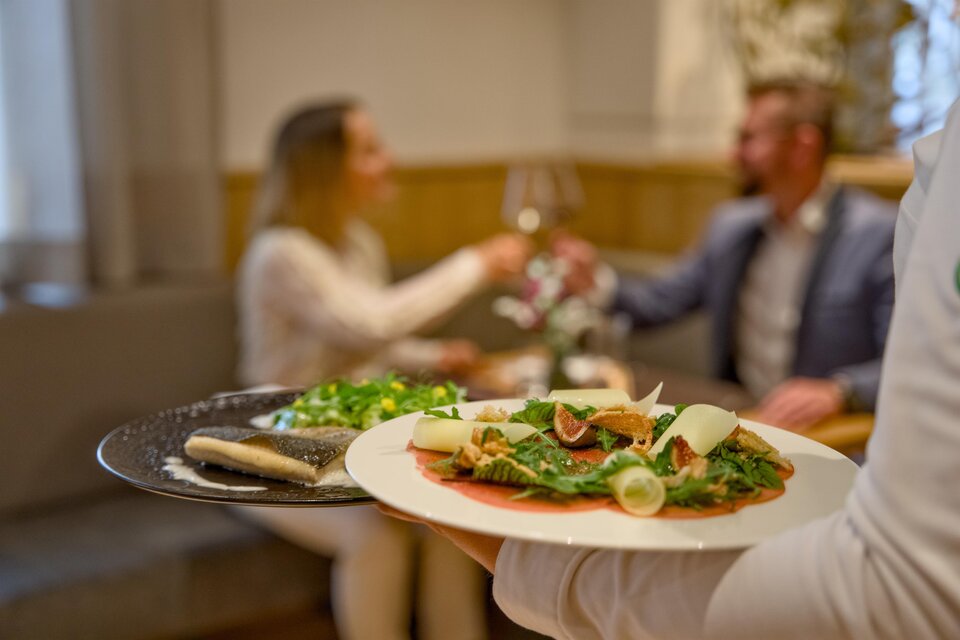 Ein gedeckter Tisch mit einem Teller voller frischem Salat und einer Hauptspeise. Im Hintergrund stoßen ein Paar mit Weingläsern an. | © Der Schrenk