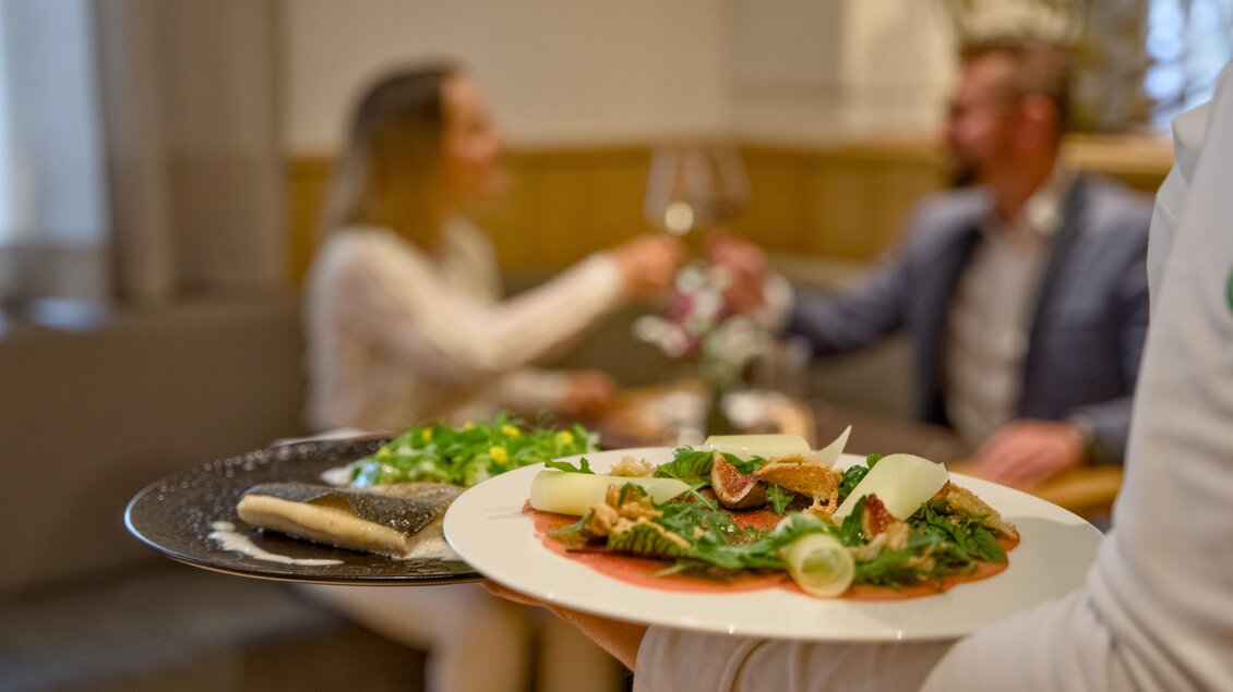 Ein gedeckter Tisch mit einem Teller voller frischem Salat und einer Hauptspeise. Im Hintergrund stoßen ein Paar mit Weingläsern an. | © Der Schrenk