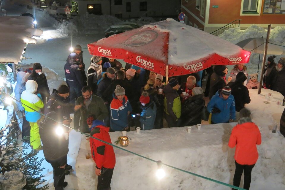 Eine große Menschenmenge versammelt sich unter einem roten Sonnenschirm im Schnee. Die Szene spielt sich bei Nacht in einer winterlichen Umgebung ab. | © Bergrettung