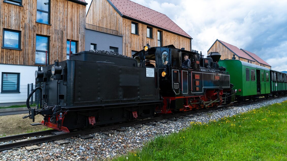 Eine dampfbetriebene Lokomotive steht neben einem grünen Zug in der Nähe moderner Gebäude. Im Hintergrund sind grüne Wiesen und ein bewölkter Himmel zu sehen. | © Berger Chris