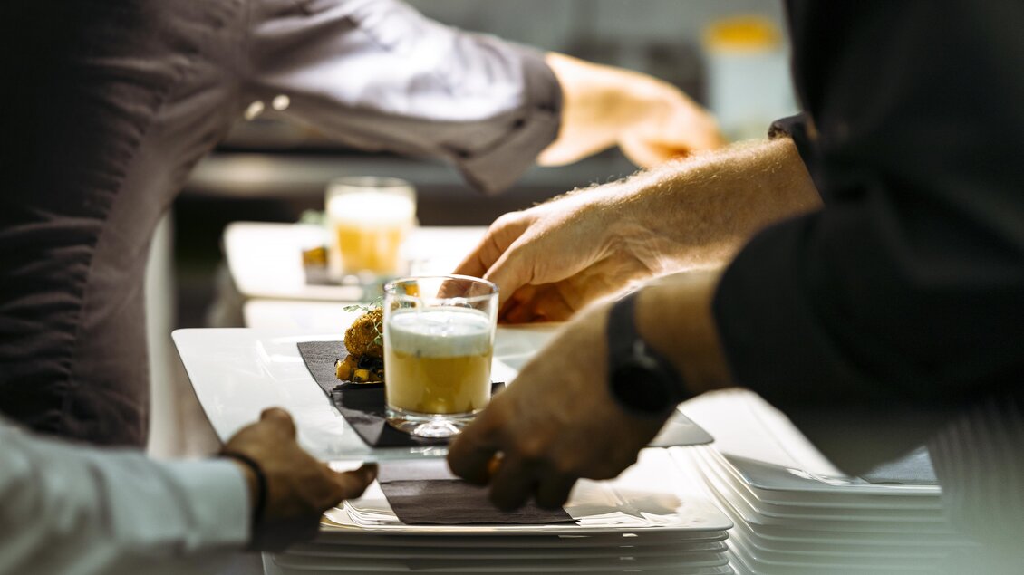 Ein geschäftiges Restaurant, in dem Teller mit Speisen und Getränken serviert werden. Die Hände von Köchen sind beim Anrichten von Gerichten zu sehen. | © Vulkanlandhotel Legenstein