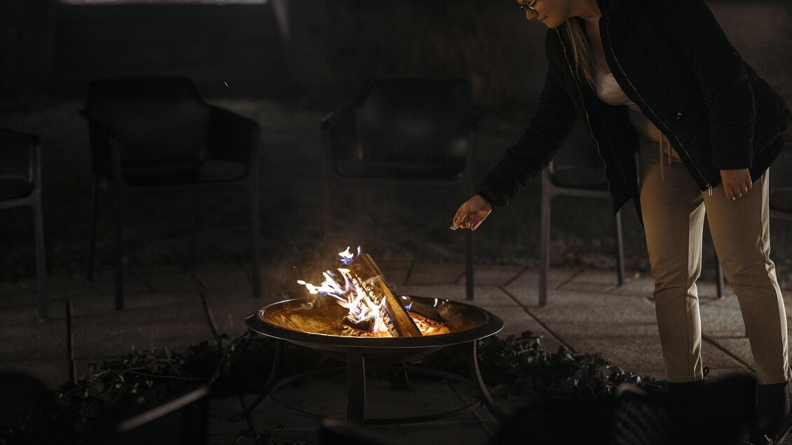 Eine Frau steht vor einem Lagerfeuer und fügt Holz hinzu. Um sie herum stehen Stühle in der Dunkelheit. | © Vulkanlandhotel Legenstein