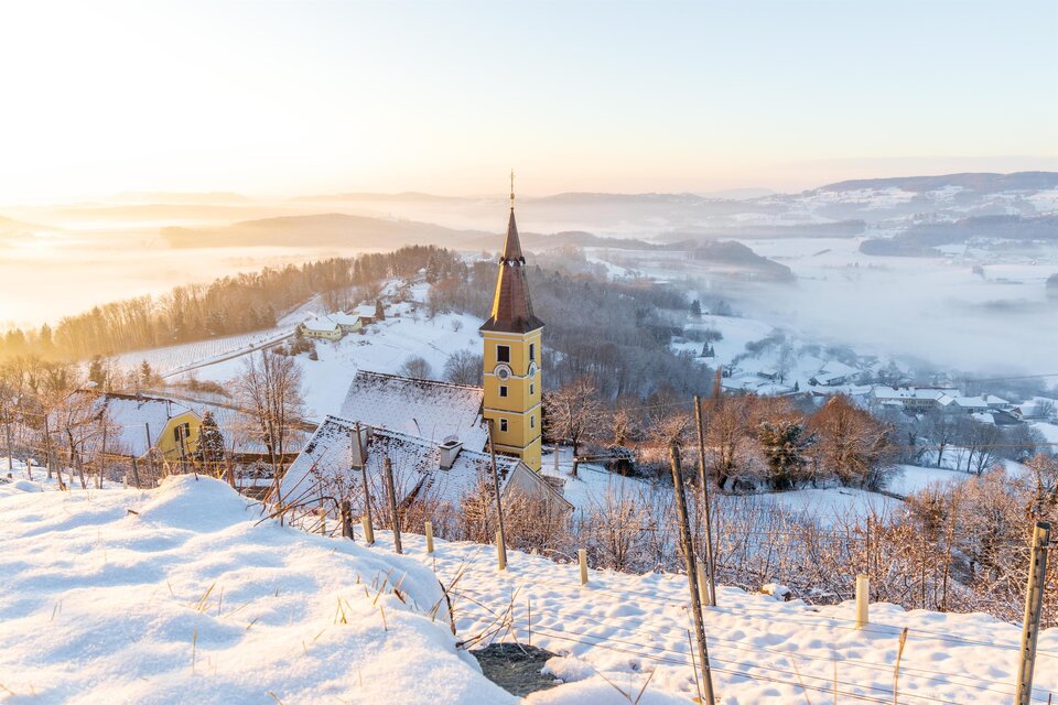 Eine verschneite Landschaft mit sanften Hügeln und einem kleinen, gelben Kirchturm. Der Sonnenaufgang sorgt für eine malerische, neblige Stimmung. | © Sebastian Friedl