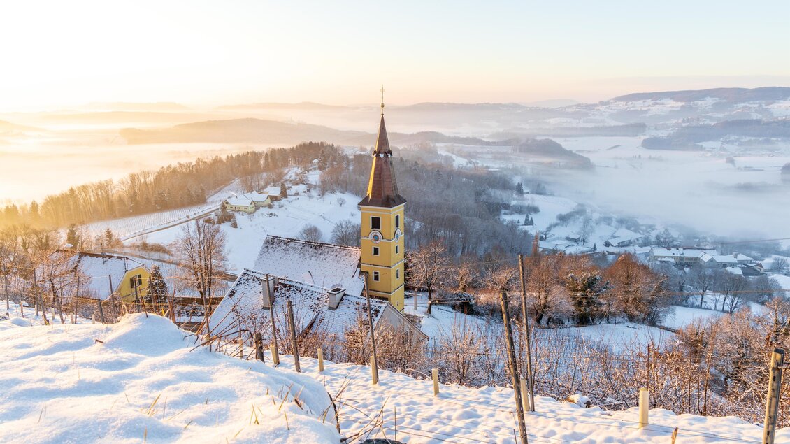 Eine verschneite Landschaft mit sanften Hügeln und einem kleinen, gelben Kirchturm. Der Sonnenaufgang sorgt für eine malerische, neblige Stimmung. | © Sebastian Friedl