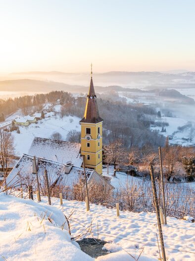 Eine verschneite Landschaft mit sanften Hügeln und einem kleinen, gelben Kirchturm. Der Sonnenaufgang sorgt für eine malerische, neblige Stimmung. | © Sebastian Friedl