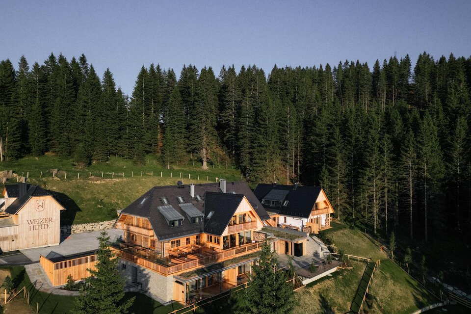 Ein gemütliches Holzhaus inmitten von hohen Tannenbäumen. Die Umgebung ist grün und einladend, ideal für einen ruhigen Rückzugsort. | © Weizerhütte