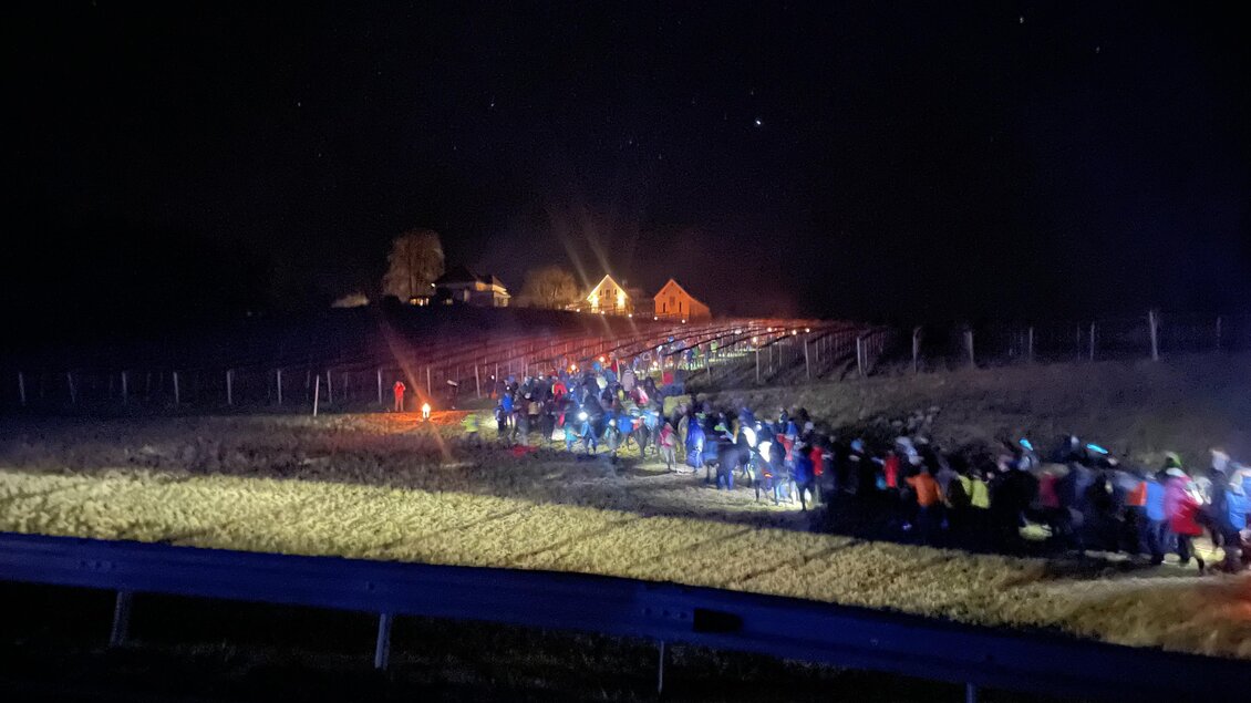 Eine große Gruppe von Menschen wandert bei Nacht entlang eines Feldes. Im Hintergrund sind beleuchtete Häuser zu sehen. | © Hannes Kofler