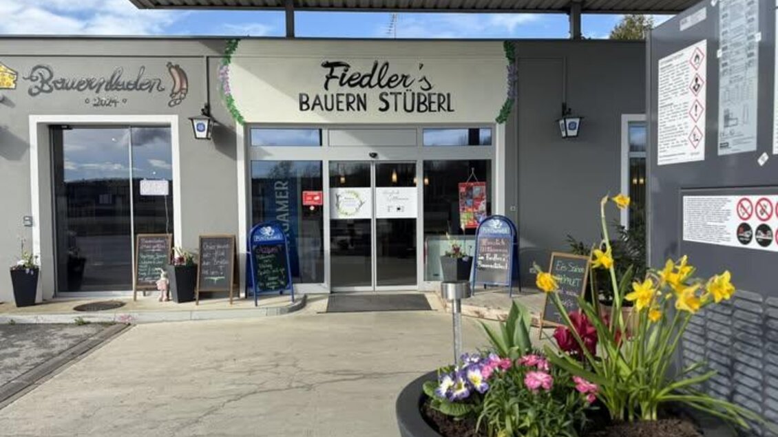 Ein einladendes Restaurant mit dem Namen "Fiedler's Bauern Stüble" und bunten Blumen vor dem Eingang. Die Fassade ist modern gestaltet, mit hellen Farben und großen Fenstern. | © Fiedler's Bauernstüberl