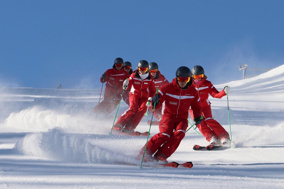 Eine Gruppe von Skifahrern in roten Anzügen fährt über eine verschneite Piste. Der klare blaue Himmel und der Schnee schaffen eine perfekte Wintersportatmosphäre. | © Hans-Peter Steiner