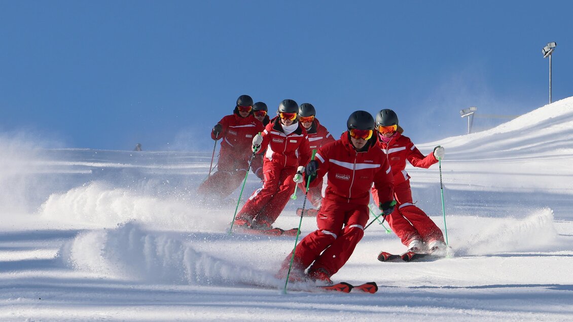 Eine Gruppe von Skifahrern in roten Anzügen fährt über eine verschneite Piste. Der klare blaue Himmel und der Schnee schaffen eine perfekte Wintersportatmosphäre. | © Hans-Peter Steiner