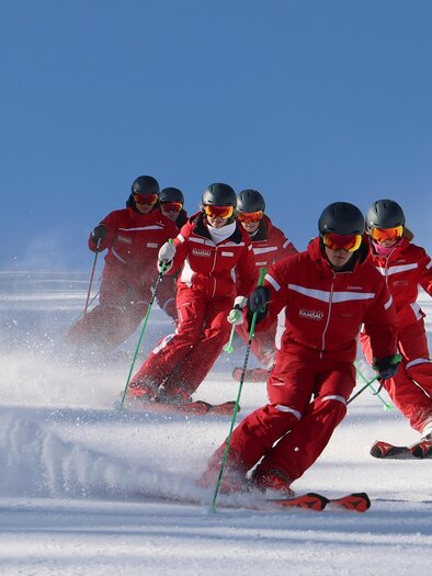 Eine Gruppe von Skifahrern in roten Anzügen fährt über eine verschneite Piste. Der klare blaue Himmel und der Schnee schaffen eine perfekte Wintersportatmosphäre. | © Hans-Peter Steiner