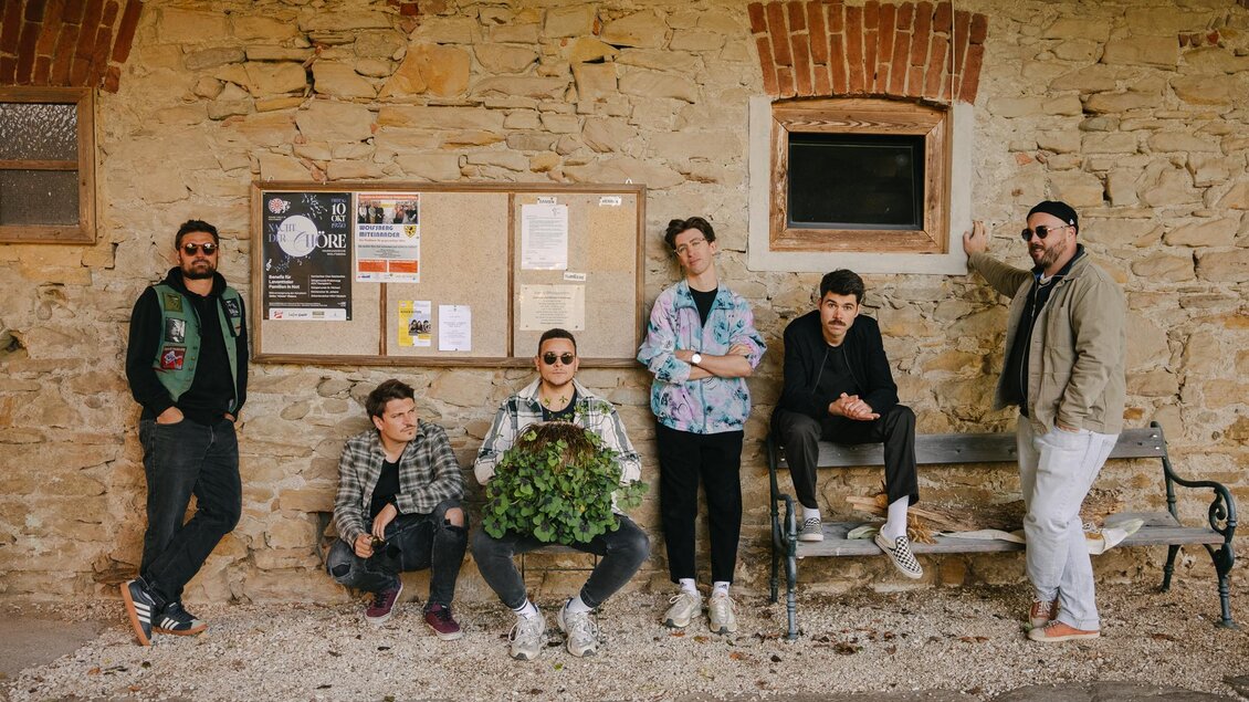 Sechs Personen stehen lässig vor einer Steinmauer mit einer Informationswand. Sie tragen modische Kleidung und posieren harmonisch zusammen. | © Fabian Flug