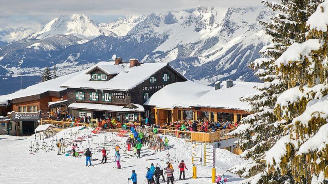 Ein Skigebiet mit einem gemütlichen Restaurant und vielen Skifahrern. Die umliegenden Berge sind schneebedeckt und der Himmel ist klar.