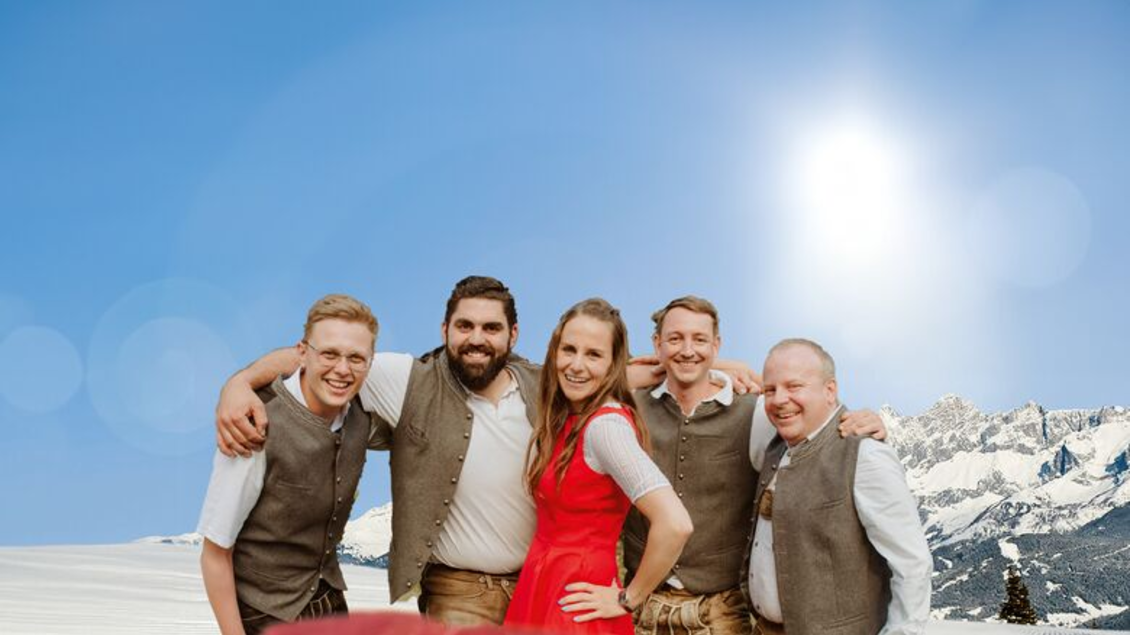 Eine Gruppe von fünf Personen steht lächelnd vor einer schneebedeckten Berglandschaft. Die Frau trägt ein rotes Kleid, während die Männer traditionelle alpine Kleidung tragen.