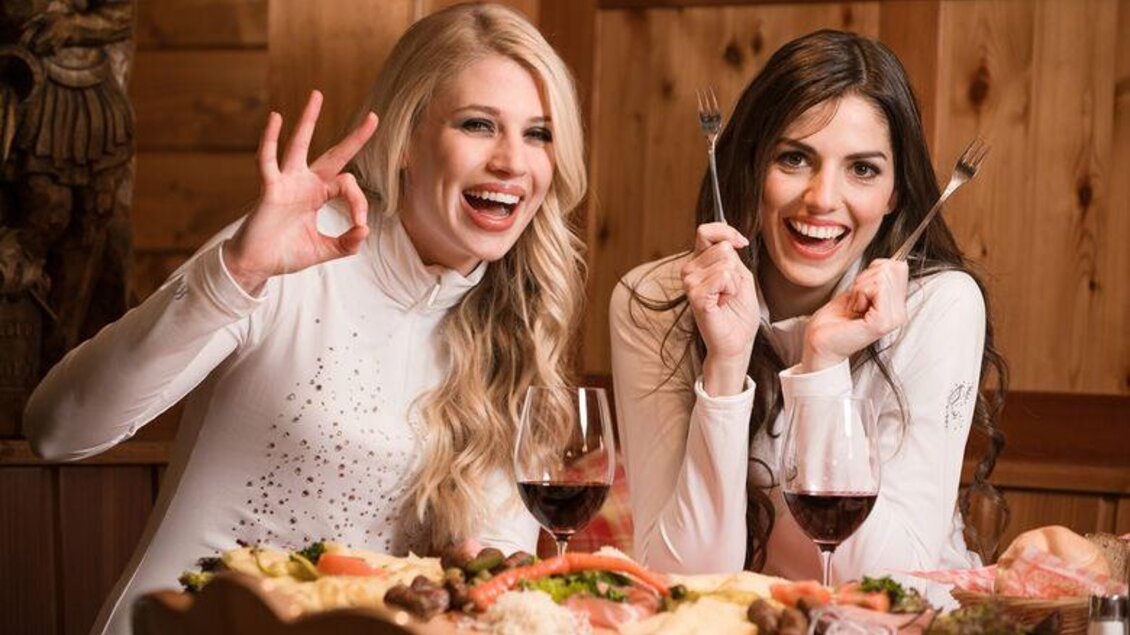 Zwei Frauen sitzen an einem Tisch und genießen ein reichhaltiges Essen. Sie lächeln und halten Gabeln in der Hand, während sie auf eine Kamera posieren.