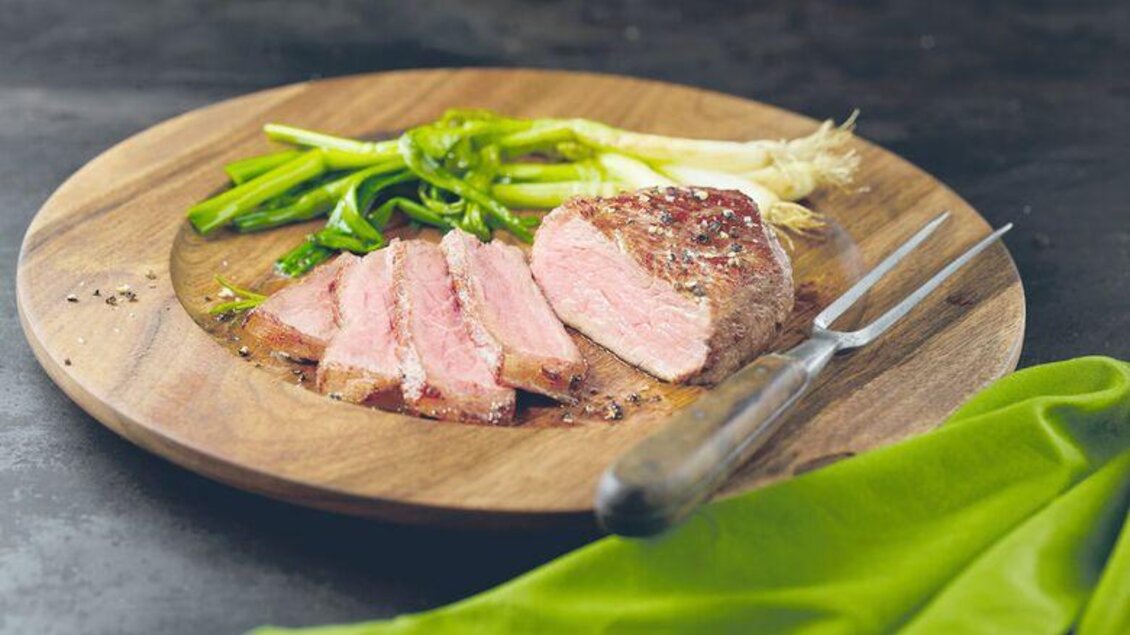 Ein Teller mit in Scheiben geschnittenem, saftigem Fleisch und frischen Frühlingszwiebeln. Daneben liegt eine grüne Serviette.