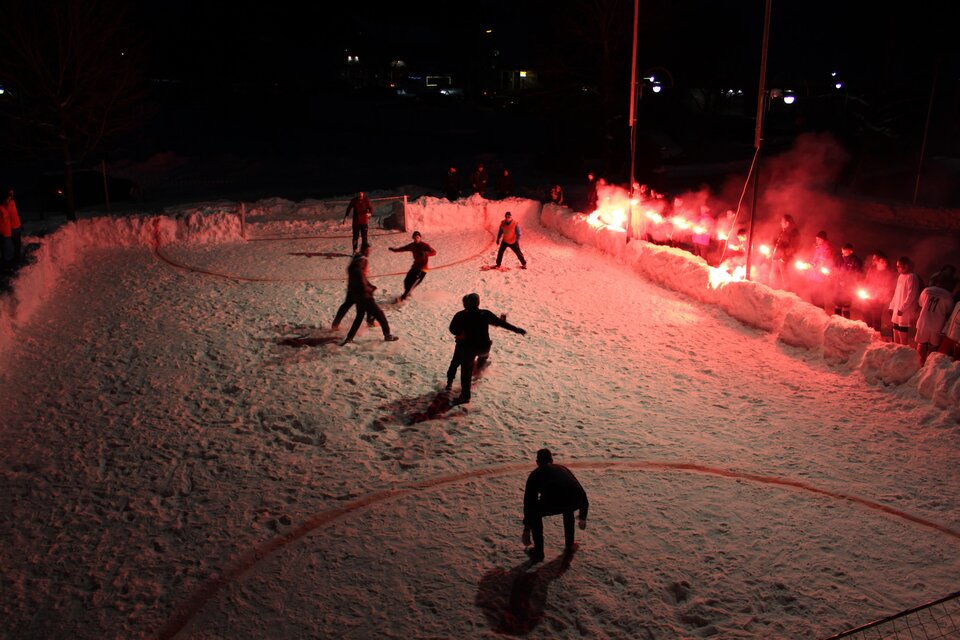 Eine Eishockeyspiel auf einem verschneiten Feld in der Nacht. Rote Bengalos erhellen die Szene und schaffen eine festliche Atmosphäre. | © Simon Treiber