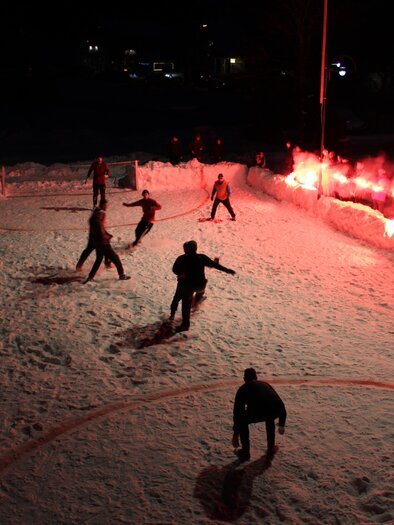 A hockey game on a snowy field at night. Red flares illuminate the scene and create a festive atmosphere. | © Simon Treiber