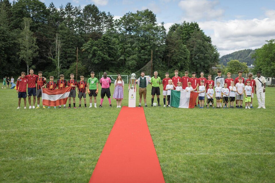 Ein Fußballspiel mit zwei Mannschaften auf einem grünen Platz. Die Spieler stehen auf einem roten Teppich, umgeben von Bäumen und unter einem teils bewölkten Himmel. | © @Martin Benedikt
