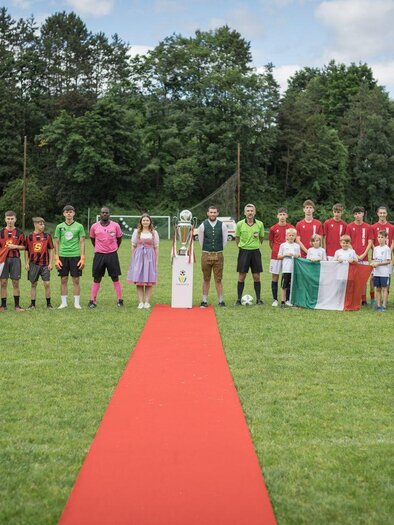 Ein Fußballspiel mit zwei Mannschaften auf einem grünen Platz. Die Spieler stehen auf einem roten Teppich, umgeben von Bäumen und unter einem teils bewölkten Himmel. | © @Martin Benedikt