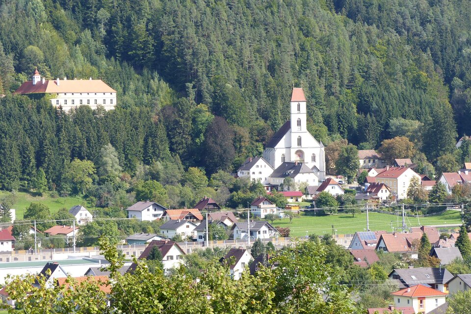 Eine malerische Landschaft mit einem kleinen Dorf und einer Kirche im Vordergrund. Im Hintergrund sind Wälder und Hügel zu sehen.