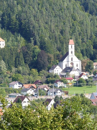 Eine malerische Landschaft mit einem kleinen Dorf und einer Kirche im Vordergrund. Im Hintergrund sind Wälder und Hügel zu sehen.