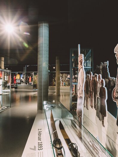 A modern museum with exhibits in display cases and murals of people. The lighting creates an inviting atmosphere. | © nicoleseiser.at