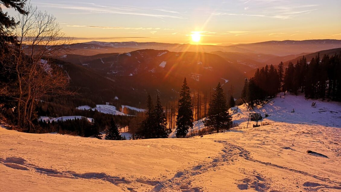 Eine verschneite Landschaft mit Bergen im Hintergrund. Die Sonne geht hinter den Hügeln auf und färbt den Himmel orange. | © steirischwandern.at