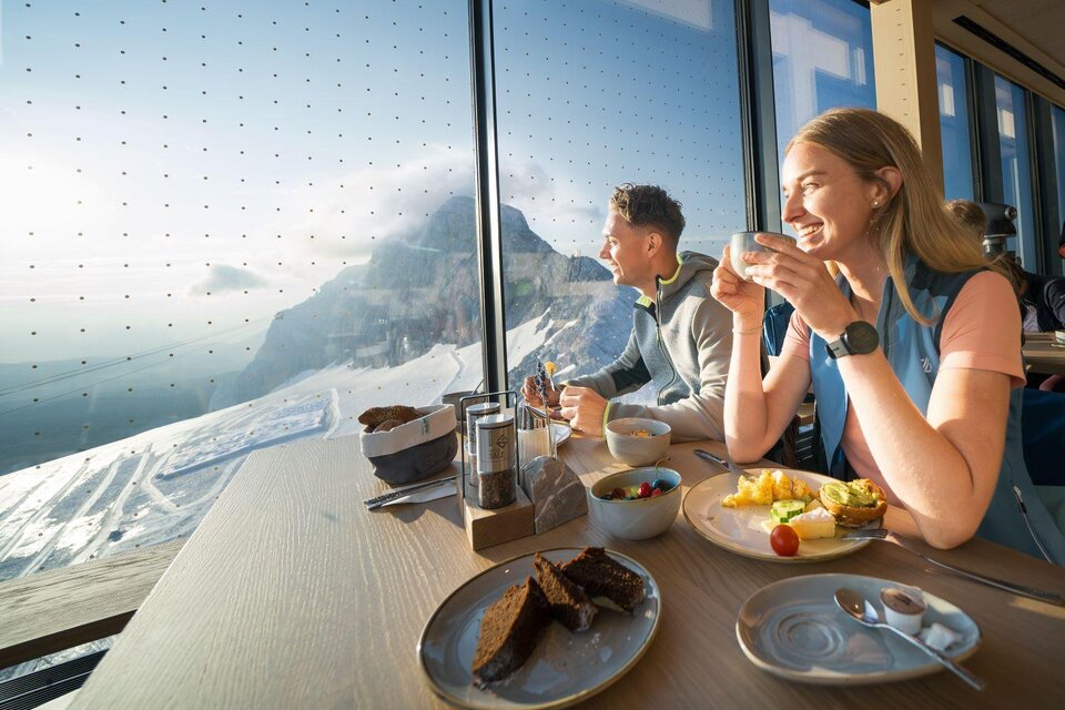 Ein Pärchen sitzt an einem Tisch mit Blick auf die Berge. Sie genießen eine Mahlzeit und trinken Kaffee, während Sonnenlicht durch große Fenster strömt. | © Josh Absenger