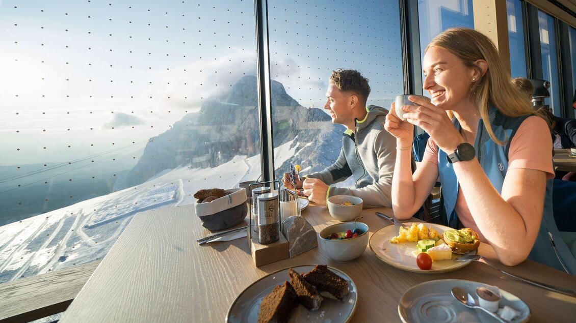 Ein Pärchen sitzt an einem Tisch mit Blick auf die Berge. Sie genießen eine Mahlzeit und trinken Kaffee, während Sonnenlicht durch große Fenster strömt. | © Josh Absenger
