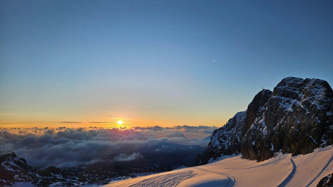 Eine malerische Schneelandschaft mit Wolken und einem Sonnenuntergang am Horizont. Im Vordergrund sind Spuren im Schnee und eine große Felsformation zu sehen. | © Pension Geierberg