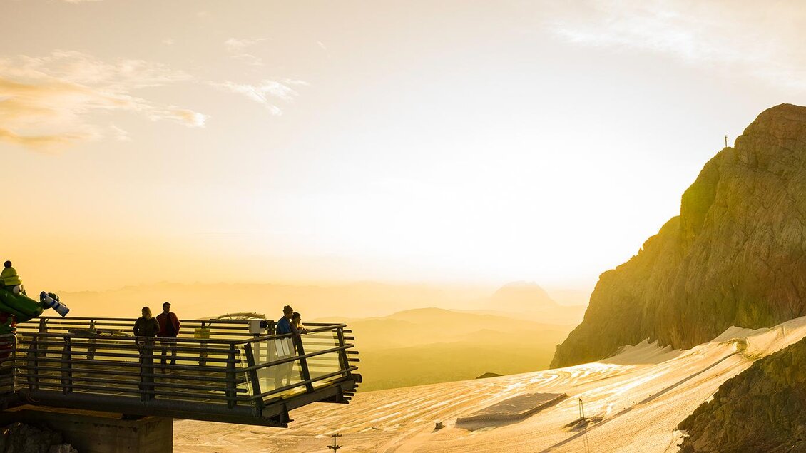Eine Aussichtplattform mit mehreren Personen bietet einen Blick auf eine beeindruckende Berglandschaft. Im Hintergrund leuchtet die untergehende Sonne am Horizont. | © Johannes Absenger
