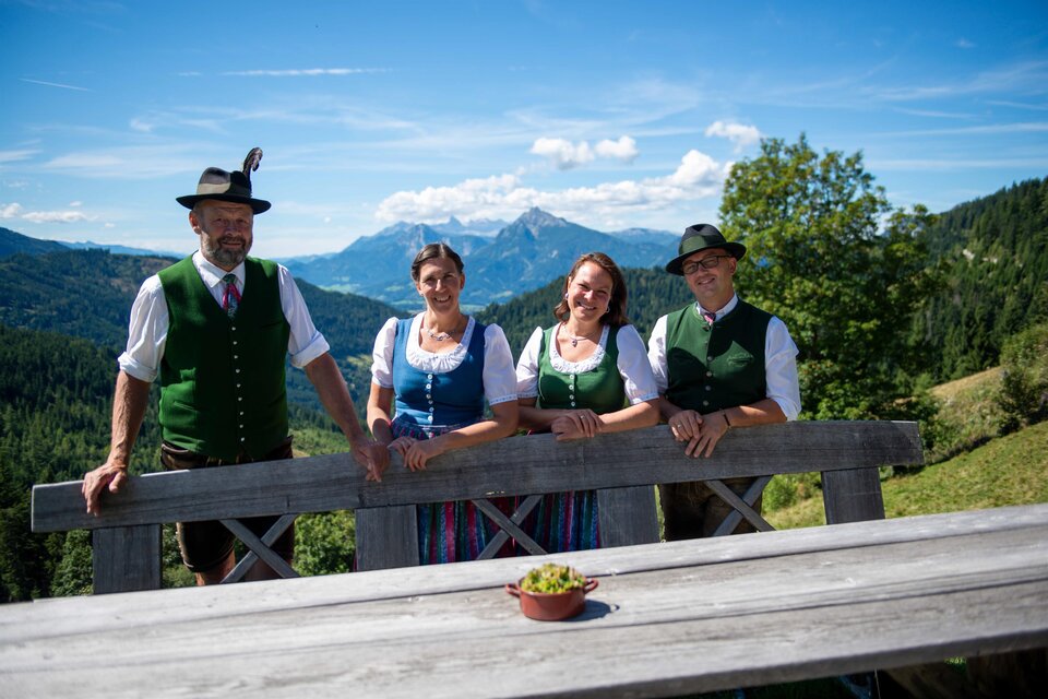 Vier Personen in Tracht (Dirndl und grüne Westen mit Hüten) stehen an einem Holzzaun auf einer Almterrasse; im Hintergrund Bergpanorama, Wälder und blauer Himmel mit Wolken. | © Hans Johann Danklmayer