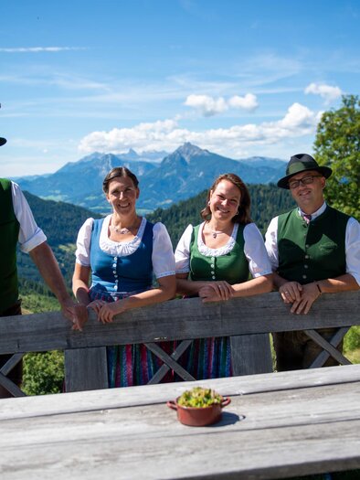 Vier Personen in Tracht (Dirndl und grüne Westen mit Hüten) stehen an einem Holzzaun auf einer Almterrasse; im Hintergrund Bergpanorama, Wälder und blauer Himmel mit Wolken. | © Hans Johann Danklmayer
