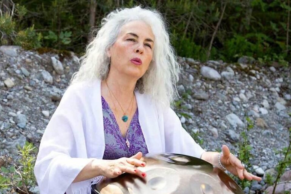 Eine Frau mit grauen Haaren spielt auf einem Handpan im Freien. Der Hintergrund zeigt einen steinigen Untergrund und grünes Laub. | © AndersOrt Haus der Frauen