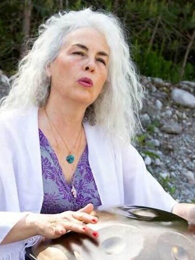 A woman with gray hair is playing a handpan outdoors. The background shows a rocky ground and green foliage. | © AndersOrt Haus der Frauen