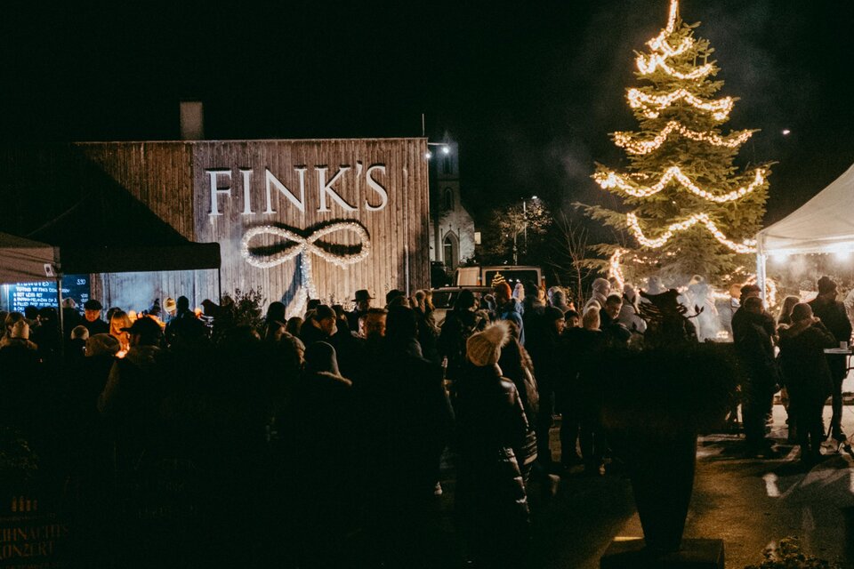 Eine festliche Veranstaltung bei Nacht mit vielen Menschen. Ein geschmückter Weihnachtsbaum leuchtet vor dem Gebäude von FINK'S. | © Haberl Fink