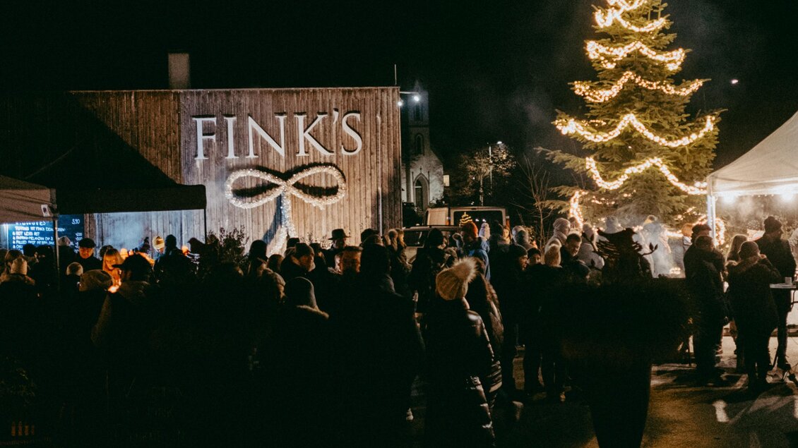 Eine festliche Veranstaltung bei Nacht mit vielen Menschen. Ein geschmückter Weihnachtsbaum leuchtet vor dem Gebäude von FINK'S. | © Haberl Fink